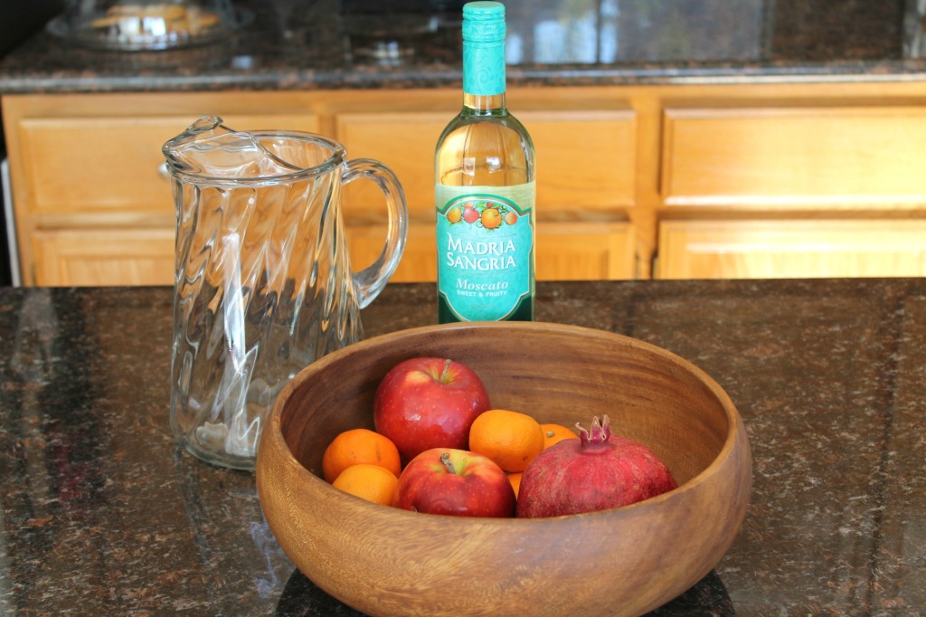 A bottle of Moscato wine, a large glass pitcher, and a bowl of cuties, apples, and a pomegranate on a kitchen counter.