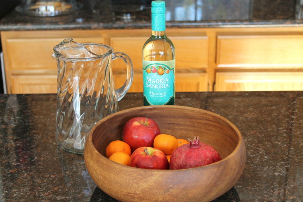 How to make a basic white wine sangria via SpanglishSpoon.com A bottle of Moscato wine, a large glass pitcher, and a bowl of cuties, apples, and a pomegranate on a kitchen counter.