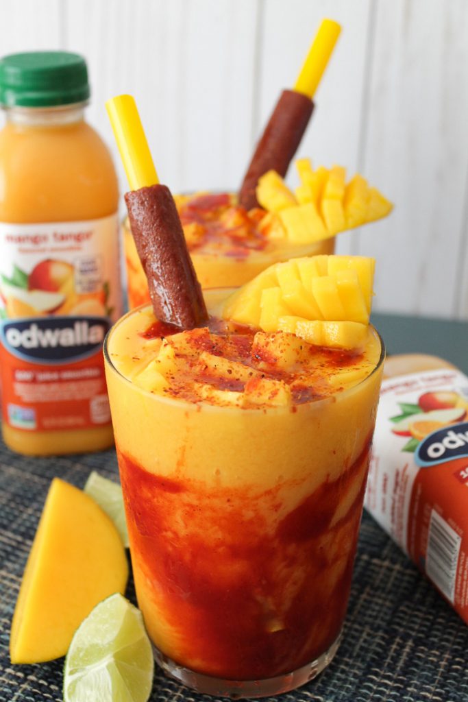 Mangonada smoothie in glass cups on a blue colored surface with two Odwalla Mango Tango drinks, lime wedges, and a mango slice on the table.