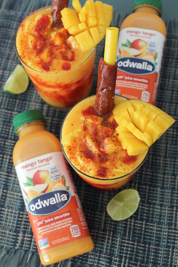 Two mangonada smoothies in glass cups on a blue colored surface with two Odwalla Mango Tango drinks, lime wedges, and a mango slice on the table.