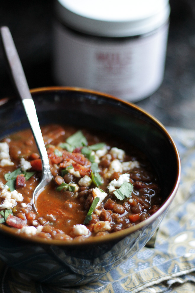 Dark blue and brown bowl with lentil soup, topped with bacon bits, cheese, and cilantro, spoon in soup.