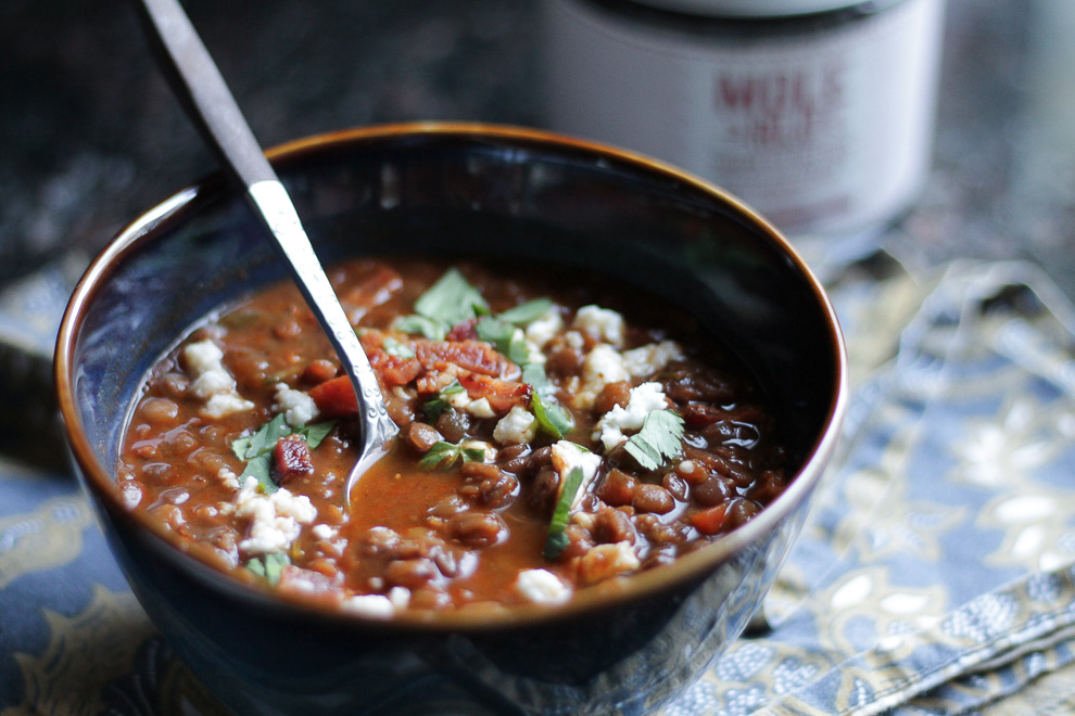 Dark blue and brown bowl with lentil soup, topped with bacon bits, cheese, and cilantro, spoon in soup.